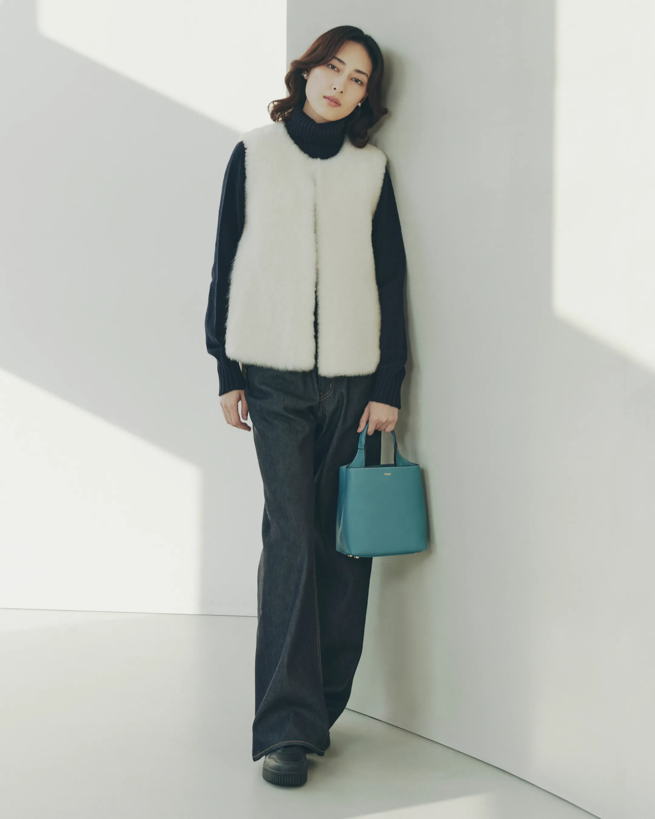 カール・ドノヒュー ジレをスカリオーネ タートルネックセーターに重ねて着用しているモデルがmica della valle ミニバッグを持ち綺麗な姿勢で立っている様子